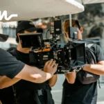 Film crew working outdoors with a RED cinema camera setup, featuring a matte box, wireless transmitter, and light modifiers. Three crew members are adjusting the equipment and managing lighting.