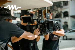 Film crew working outdoors with a RED cinema camera setup, featuring a matte box, wireless transmitter, and light modifiers. Three crew members are adjusting the equipment and managing lighting.